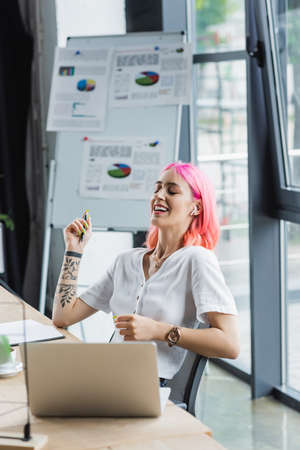 happy businesswoman in earphone holding pen and laughing near laptopの写真素材