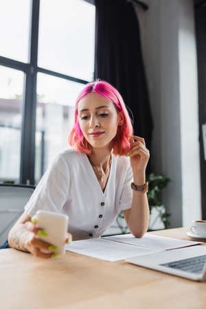 smiling businesswoman with pink hair looking at smartphone near laptopの写真素材