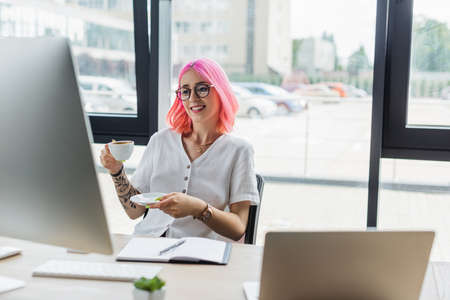 happy businesswoman with pink hair holding cup of coffee and looking at computer monitorの写真素材