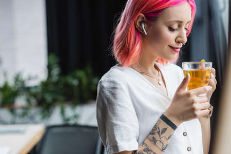 pleased businesswoman with pink hair and earphone holding cup of tea in office