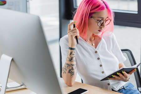 tattooed businesswoman with pink hair holding pen and notebookの写真素材
