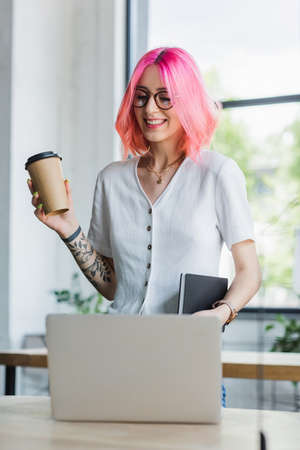 happy pierced manager with pink hair holding paper cup and looking at laptopの写真素材