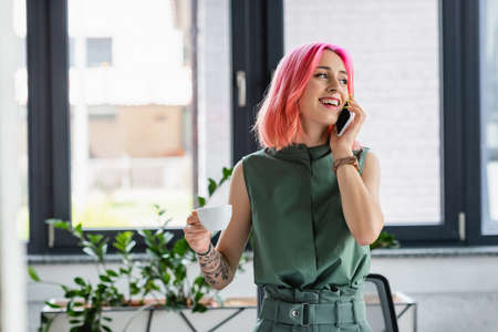 cheerful businesswoman with pink hair and piercing holding cup of coffee while talking on smartphone in officeの写真素材
