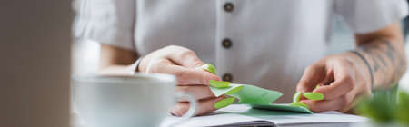 cropped view of tattooed businesswoman holding sticky note near notebook and blurred cup, bannerの写真素材