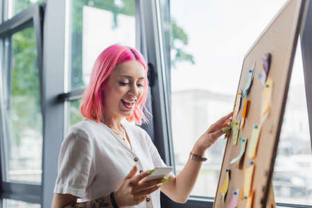 happy businesswoman with pink hair holding smartphone and pointing at board with sticky notes