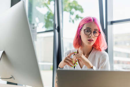 businesswoman with pink hair holding pen and looking at laptop near computer monitorの写真素材