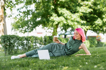 smiling young woman with pink hair holding paper cup near laptop on grassの写真素材