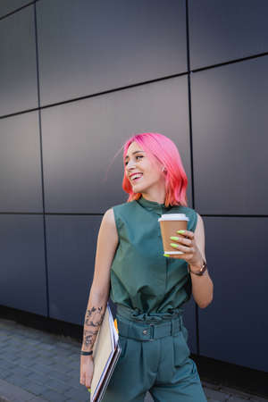 smiling businesswoman with pink hair and earphone holding coffee to go and folders
