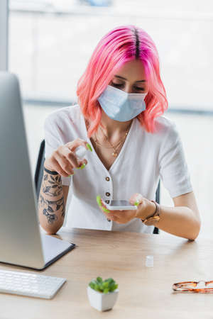 young businesswoman in medical mask spraying sanitizer on smartphone in officeの写真素材
