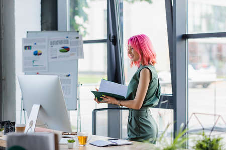 joyful businesswoman with pink hair holding folder and looking at computer monitorの写真素材