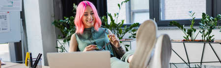 happy businesswoman with pink hair holding eyeglasses while looking at laptop, banner