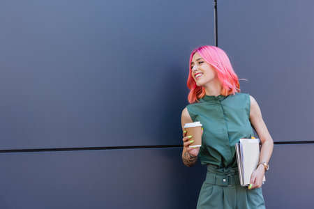 tattooed and smiling businesswoman with pink hair holding folder and coffee to go outside