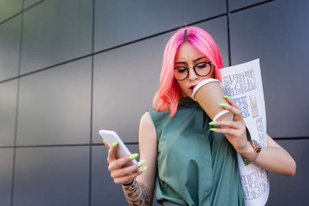 tattooed businesswoman with pink hair holding smartphone and paper cup