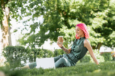 cheerful young woman with pink hair holding paper cup near laptop on grass