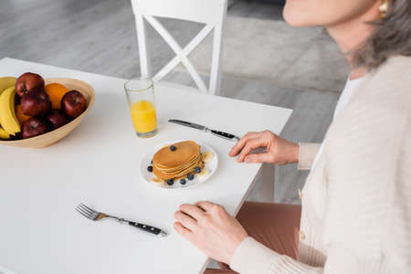 Cropped view of mature woman sitting near pancakes, orange juice and fruits in kitchen