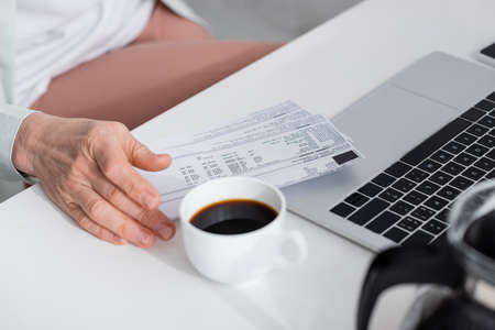 Cropped view of mature woman holding bills near coffee and laptop on table