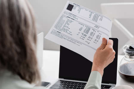 Paper with bills in hand of mature woman near laptops and coffee at home
