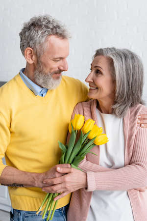Smiling woman holding flowers near bearded husband at homeの写真素材