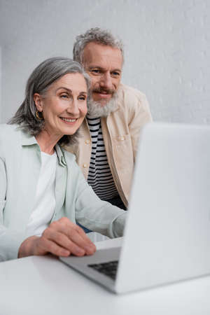 Positive freelancer using laptop near middle aged husband at home