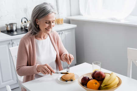 Smiling woman cutting pancakes near ripe fruits in kitchenの写真素材