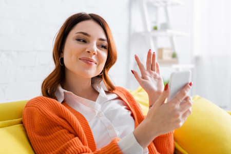 smiling woman waving hand during video call on cellphone at home