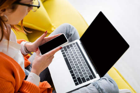 cropped view of blurred woman holding laptop with blank screen while sitting on couch with laptop