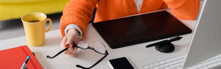 partial view of retoucher holding eyeglasses near graphic tablet, smartphone with blank screen and...