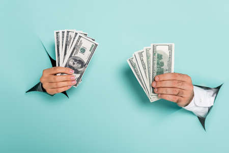 cropped view of man and woman holding dollar banknotes through holes in paper wall on blue