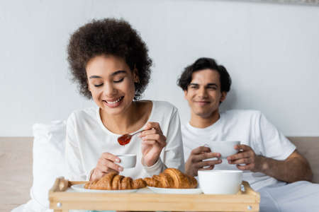 cheerful african american woman holding spoon with jam near croissants and blurred boyfriend with...