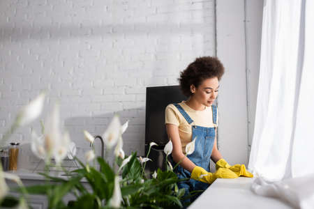 curly african american woman holding rag near window sill and plants in kitchen