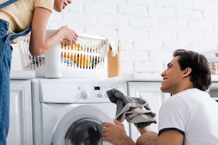 smiling man loading washing machine near cheerful african american girlfriend with basket of dirty...