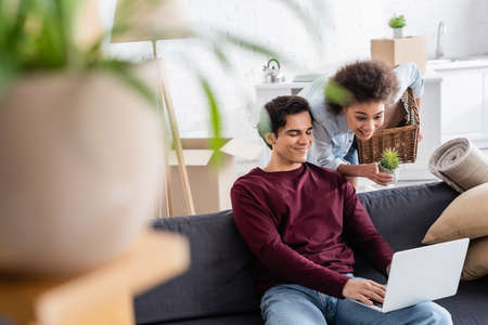 cheerful african american woman holding basket and plant near smiling boyfriend using laptop