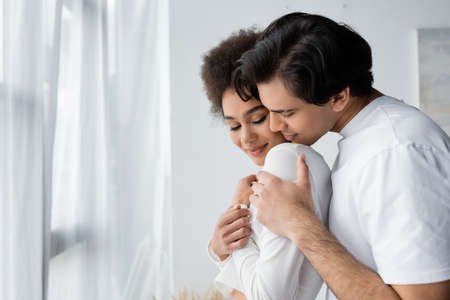 young man hugging smiling african american girlfriend at homeの写真素材