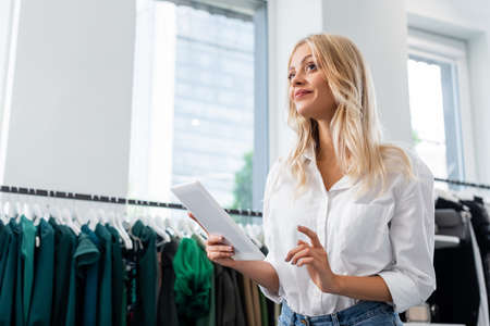 pleased sales assistant in white shirt holding digital tablet in clothing boutiqueの写真素材