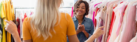 blurred woman pointing with finger at clothing near cheerful african american shop assistant in...