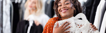 joyful african american woman holding hanger with white dress near blurred client in boutique, ...