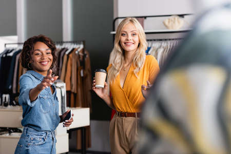 happy woman holding paper cup while african american shop assistant with smartphone pointing awayの写真素材