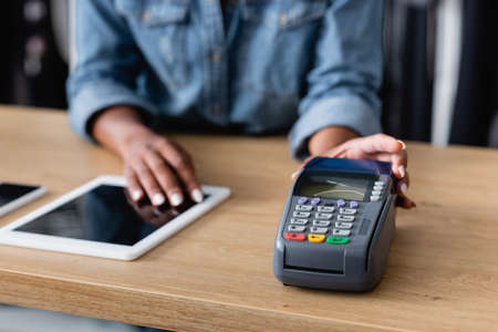 cropped view of african american saleswoman holding payment terminal near gadgets on sales counter deskの写真素材