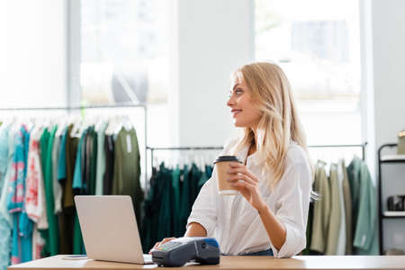happy saleswoman holding paper cup near gadgets at sales counter desk