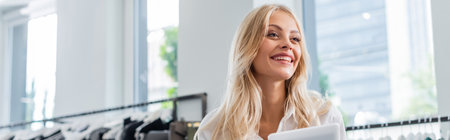 joyful sales assistant in white shirt holding digital tablet in clothing boutique, bannerの写真素材