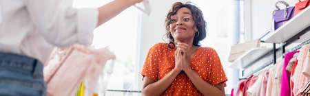 joyful african american woman looking at friend holding blurred pink dress, bannerの写真素材