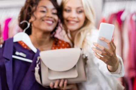 blurred blonde woman holding purse and taking selfie with cheerful african american friendの写真素材