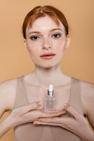Young redhead woman holding cosmetic serum and looking at camera isolated on beigeの写真素材