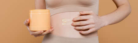 Cropped view of young woman applying cream on belly isolated on beige, banner