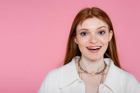 Positive freckled woman in jacket and necklaces looking at camera isolated on pink