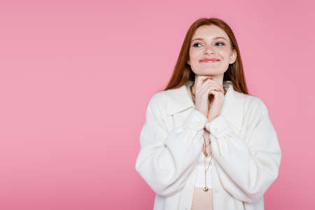Pleased red haired woman in necklaces and jacket isolated on pinkの写真素材
