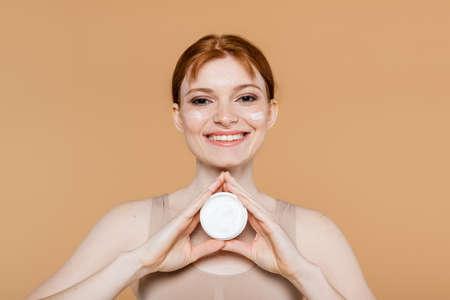Smiling redhead woman holding cosmetic cream and looking at camera isolated on beigeの写真素材