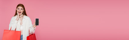 Excited redhead woman with shopping bags holding cellphone with blank screen isolated on pink,...