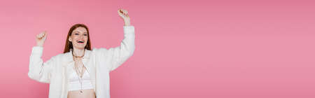 Excited red haired woman in jacket and necklaces looking at camera isolated on pink, bannerの写真素材
