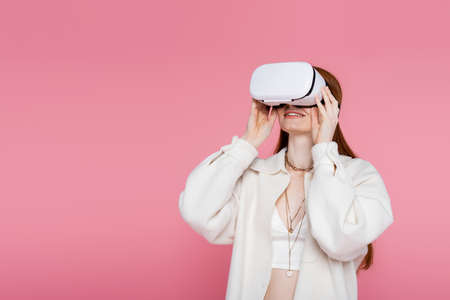 Smiling red haired woman in jacket and necklaces gaming in vr headset isolated on pinkの写真素材